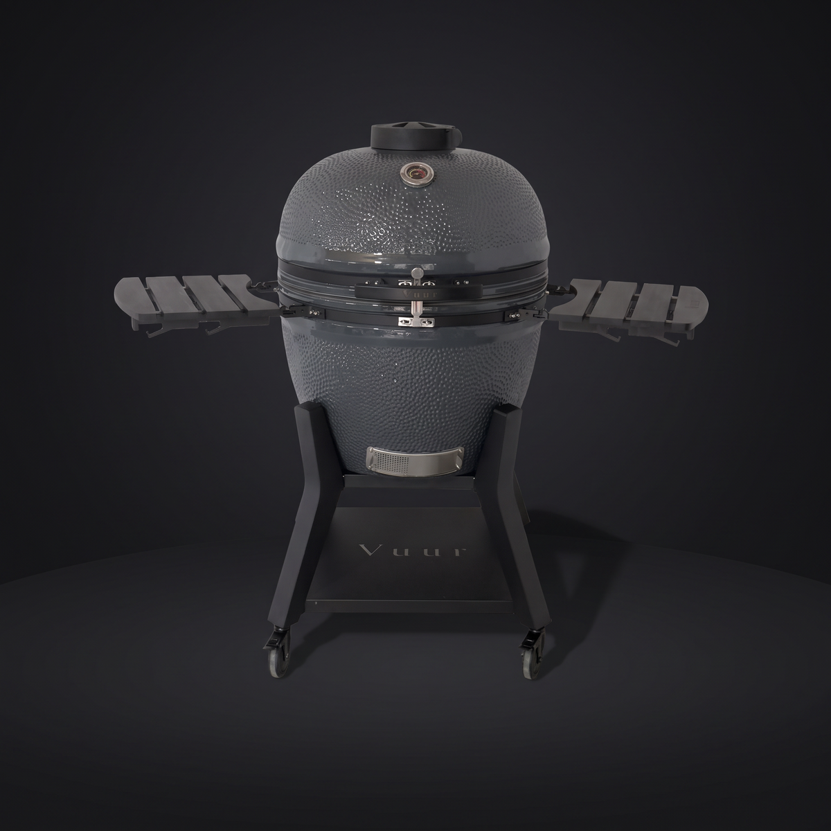 Grijze Vuur 27 kamado, gesloten, rechte voorkant, op het zwarte stalen onderstel met wielen, op een donkere achtergrond. De opklapbare zijtafels staan ingeklapt.